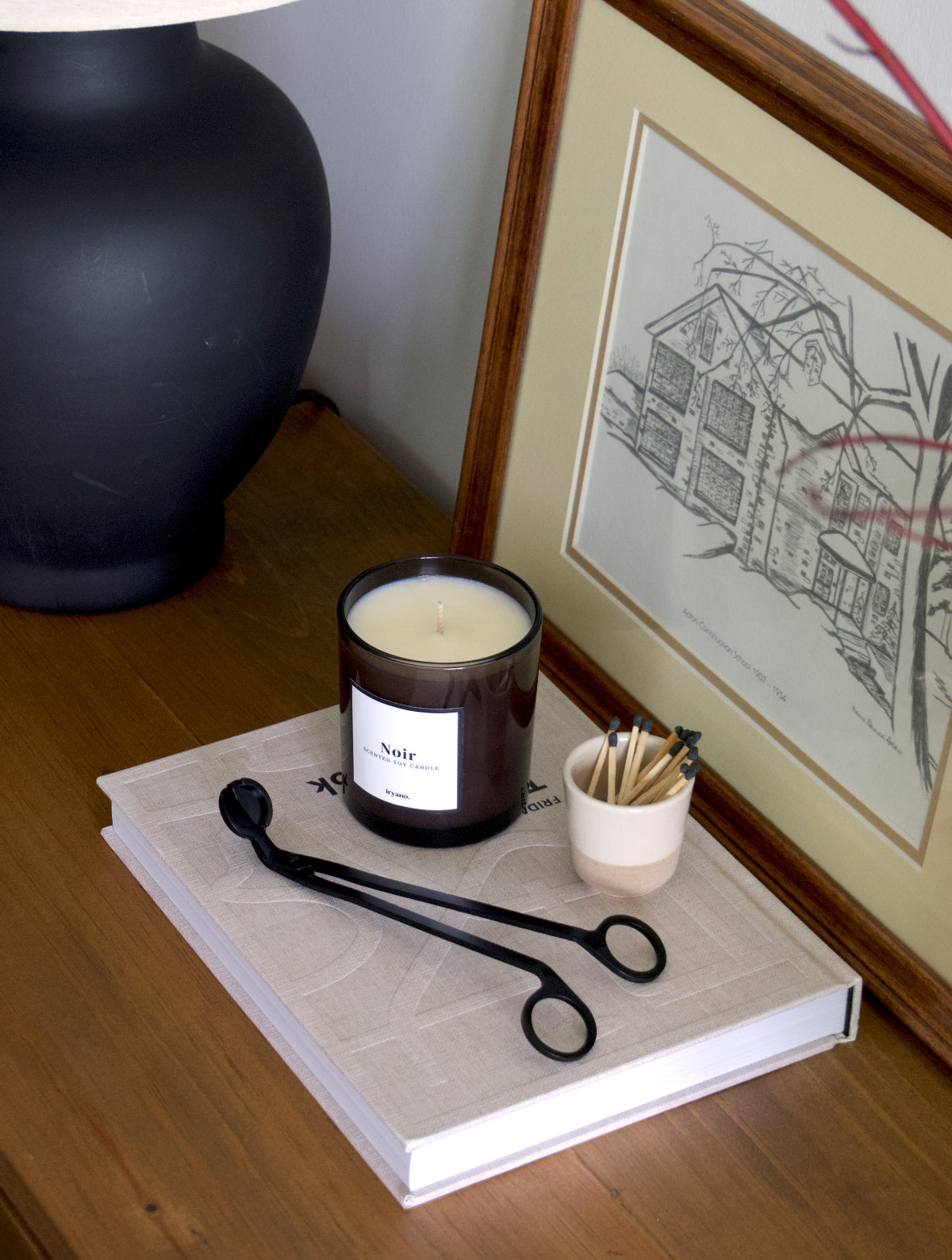 A wick trimmer and a candle in front of a picture frame and a lamp.