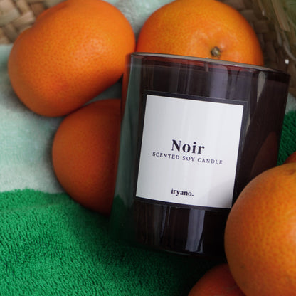 Scented soy candle labeled 'Noir' by iryano surrounded by oranges on a green surface.