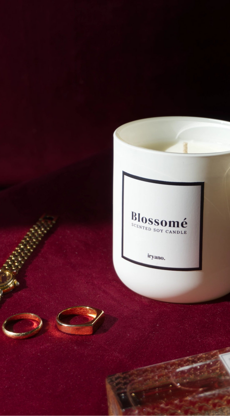 Candle labeled 'Blossomé' on a red surface with rings and a perfume bottle.