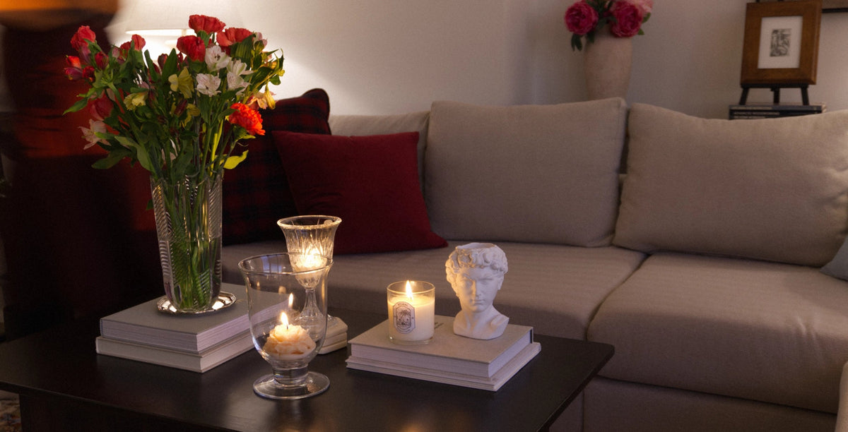 A cozy living room scene featuring a beige sofa with red accent pillows, a dark coffee table decorated with books, lit candles in glass holders, and a large vase of red and white roses.