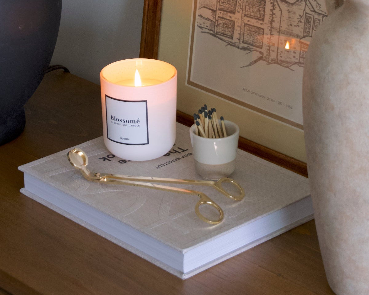 A gold wick trimmer and a candle in front of a picture frame and a lamp.