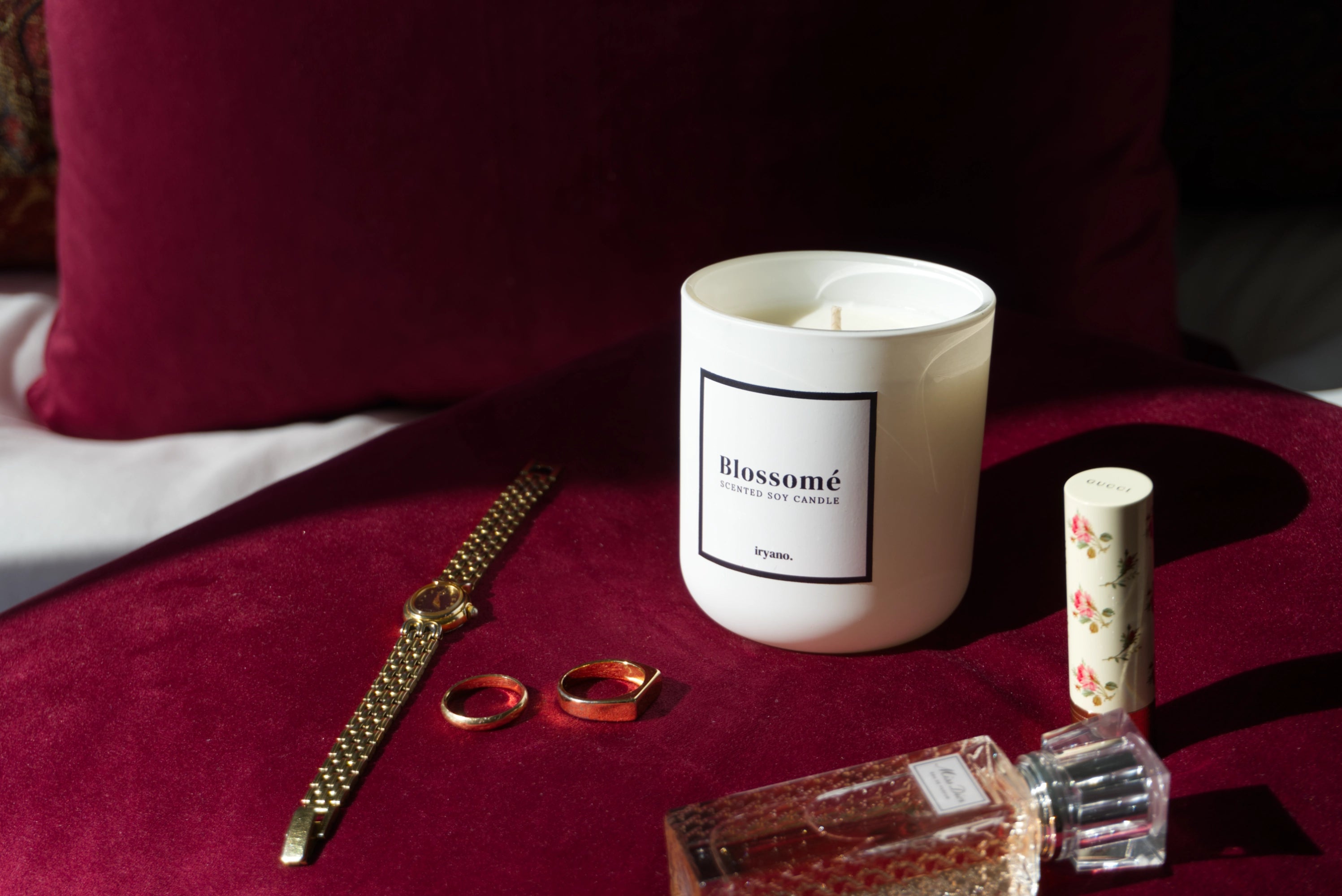 Candle labeled 'Blossomé' on a red surface with rings and a perfume bottle.