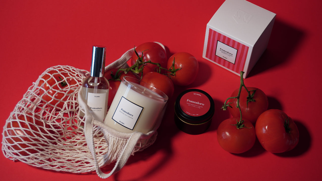 Perfume bottle, jar, and small container on a red surface with tomatoes and a mesh bag.