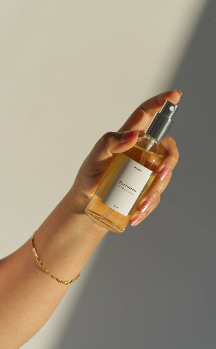 A hand holding a clear spray bottle filled with amber liquid, featuring a white label and a silver pump, set against a minimalist beige and gray background with soft diagonal shadows.