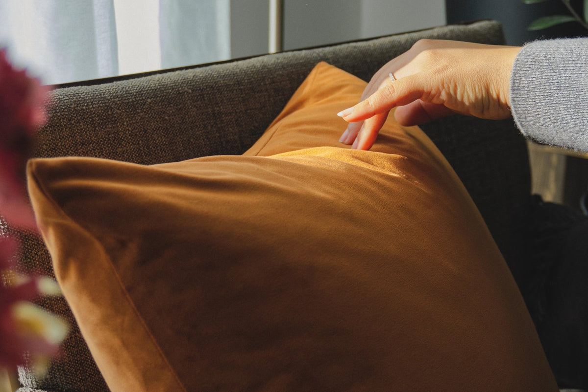 A person's hand rests on a warm camel-brown velvet throw pillow resting on a dark gray sofa in a cozy, sunlit living room setting.